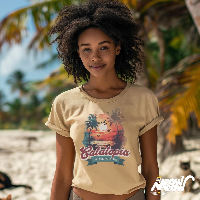 May include: A tan t-shirt with rolled sleeves, displaying a sunset scene with palm trees, birds, and the text "Catatopia Feline Paradise." The shirt is worn by a person on a beach with palm trees in the background.