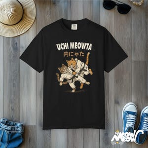 K&ouml;nnte beinhalten: Ein schwarzes Kurzarm-T-Shirt mit einer Grafik von zwei Katzen in wei&szlig;en Kampfkunst-Uniformen mit schwarzen G&uuml;rteln. Der Text UCHI MEOWTA und japanische Schriftzeichen sind in einem hellen Cremeton &uuml;ber den Katzen aufgedruckt.