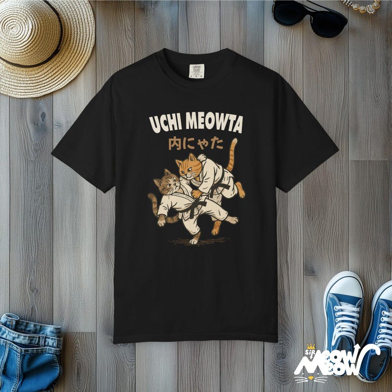 K&ouml;nnte beinhalten: Ein schwarzes Kurzarm-T-Shirt mit einer Grafik von zwei Katzen in wei&szlig;en Kampfkunst-Uniformen mit schwarzen G&uuml;rteln. Der Text UCHI MEOWTA und japanische Schriftzeichen sind in einem hellen Cremeton &uuml;ber den Katzen aufgedruckt.