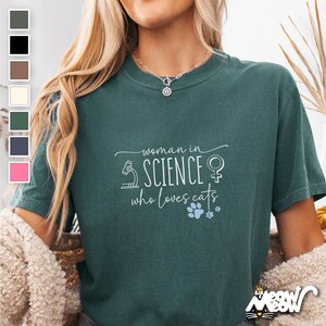 May include: A teal green t-shirt with the text "woman in science who loves cats" in white. The text is surrounded by a small microscope, a female symbol, and a paw print.