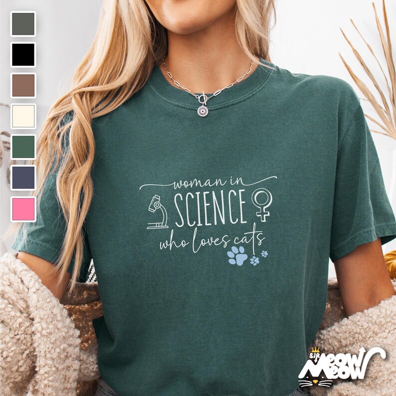 May include: A teal green t-shirt with the text "woman in science who loves cats" in white. The text is surrounded by a small microscope, a female symbol, and a paw print.