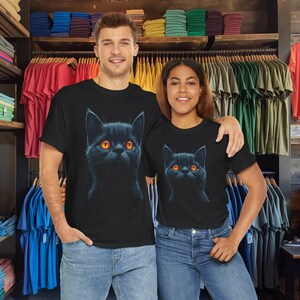 May include: Black t-shirts featuring a detailed illustration of a black cat with bright orange eyes. The cat's portrait is centered on the shirt. The models are wearing blue jeans. The background shows a clothing store with racks of colorful t-shirts.