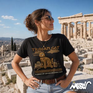 May include: A black short-sleeve t-shirt featuring a graphic of a wooden cat on wheels, resembling a Trojan horse. The text reads "TROJAN CAT" and "BEWARE OF GREEKS BEARING CATS" in a tan, blocky font, with a Greek key border design at the bottom.