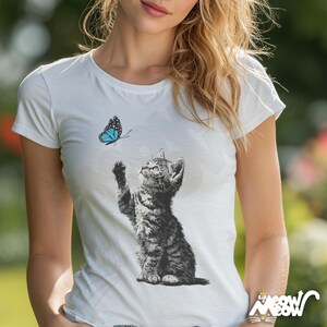 May include: White t-shirt featuring a black and white illustration of a kitten reaching for a blue butterfly. The kitten is detailed with shading, and the butterfly has blue wings with black accents. The shirt has a crew neck and short sleeves.