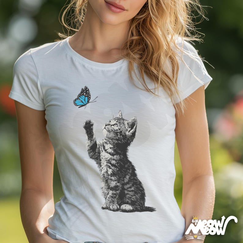 May include: White t-shirt featuring a black and white illustration of a kitten reaching for a blue butterfly. The kitten is detailed with shading, and the butterfly has blue wings with black accents. The shirt has a crew neck and short sleeves.