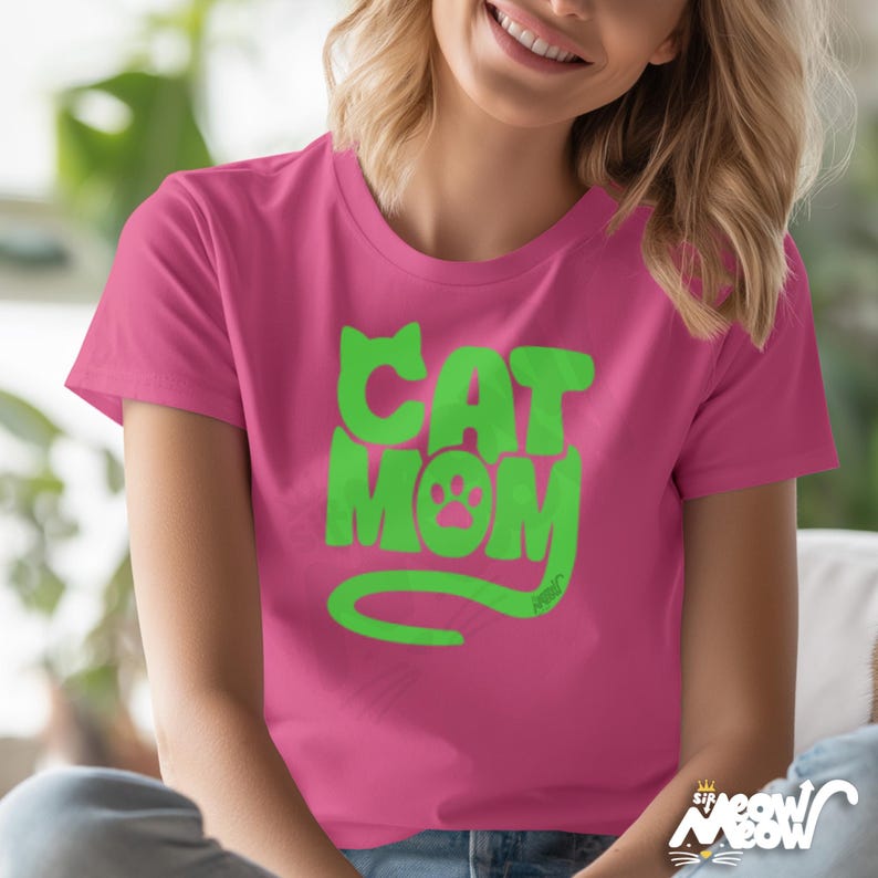 May include: A pink t-shirt with the words "CAT MOM" in bright green, featuring a paw print in the letter "O". The shirt has short sleeves and a crew neck. The design is centered on the front of the shirt.