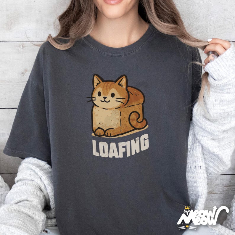 Op de afbeelding: Een donkergrijs T-shirt met een cartoonkat in de vorm van een brood. De kat is bruin en oranje met een wit gezicht en de tekst "loafing" is onder de kat gedrukt.