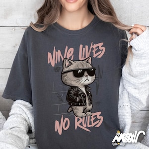 May include: Dark gray t-shirt featuring a cartoon cat wearing sunglasses and a black leather jacket. The text "NINE LIVES NO RULES" is printed above and below the cat illustration in a pink font. The shirt has a relaxed fit.