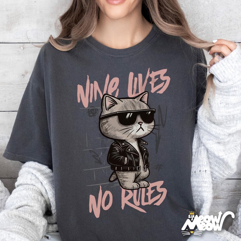 May include: Dark gray t-shirt featuring a cartoon cat wearing sunglasses and a black leather jacket. The text "NINE LIVES NO RULES" is printed above and below the cat illustration in a pink font. The shirt has a relaxed fit.