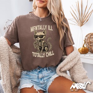 May include: A brown t-shirt featuring a cartoon skeleton cat wearing sunglasses and holding a microphone. The text "Mentally Ill Totally Chill" is printed on the shirt.