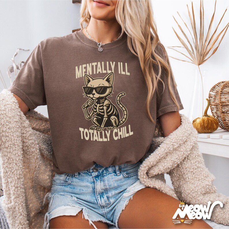 May include: A brown t-shirt featuring a cartoon skeleton cat wearing sunglasses and holding a microphone. The text "Mentally Ill Totally Chill" is printed on the shirt.