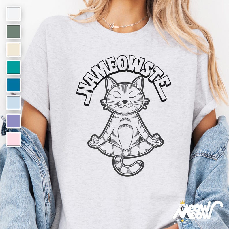 Peut inclure: Un t-shirt gris avec le texte "Nameowste" en contour noir. Une illustration en noir et blanc d'un chat assis en position de yoga est centr&eacute;e sous le texte.