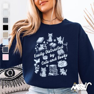 Könnte beinhalten: Ein marineblauer Pullover mit einem weißen Grafikdesign mit Katzen, Büchern und dem Text "Easily Distracted by Cats and Books".