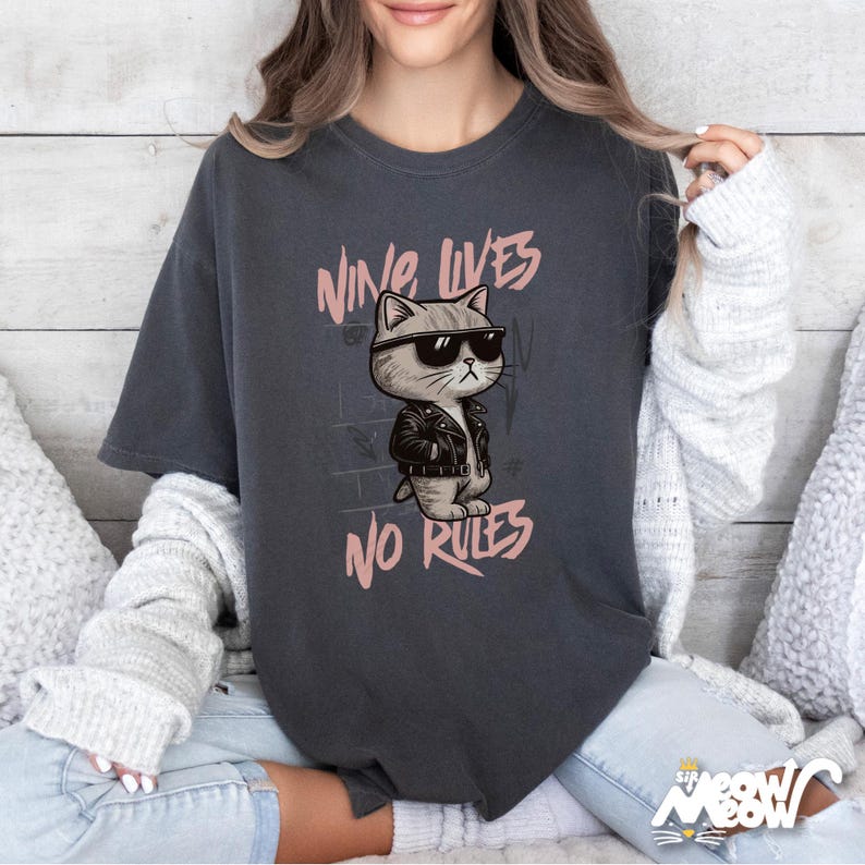 May include: Dark gray t-shirt featuring a cartoon cat wearing sunglasses and a leather jacket. The text "NINE LIVES NO RULES" is printed above and below the cat illustration in pink. The shirt has short sleeves and a relaxed fit.
