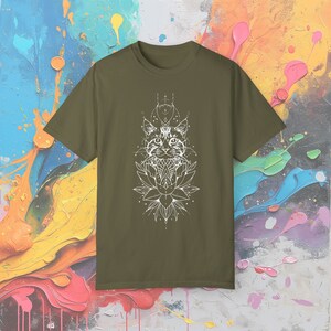 May include: Olive green t-shirt featuring a white graphic of a cat's face, a lotus flower, and geometric shapes. The design is centered on the front of the shirt. The shirt is made of a soft material.