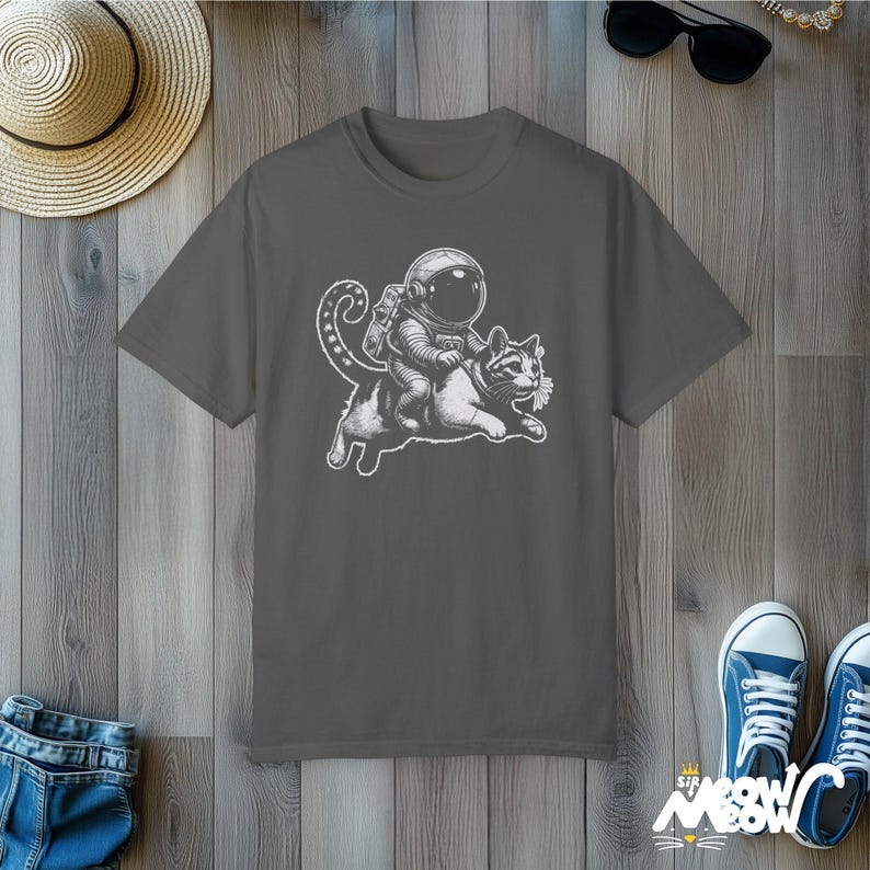 Pepper colored Comfort Colors 1717 unisex T-shirt with a humorous white graphic of an astronaut riding atop a playful cat, combining outer space and cat lover themes.