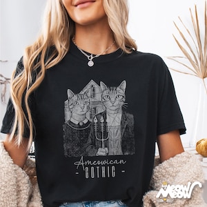 May include: Black t-shirt featuring a parody of the American Gothic painting, with two cats in place of the original figures. The text "American Gothic" is below the image. The shirt has a relaxed fit.