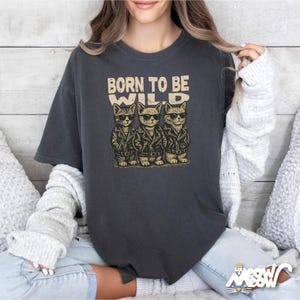 Puede incluir: Camiseta gris oscuro con un gr&aacute;fico de tres gatos con gafas de sol y chaquetas de cuero. El texto "BORN TO BE WILD" est&aacute; impreso encima de los gatos. La camiseta tiene un corte holgado.