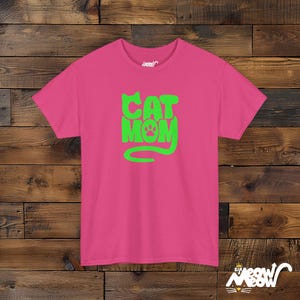 May include: A pink t-shirt with the words "CAT MOM" in bright green, featuring a paw print in the letter "O" and a curved tail. The shirt is made of a soft material and is suitable for casual wear.
