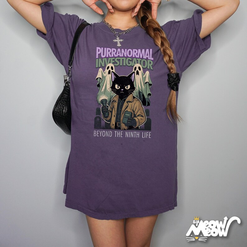 May include: A purple t-shirt featuring a cartoon cat dressed as a paranormal investigator, with the text "PURRANORMAL INVESTIGATOR" and "BEYOND THE NINTH LIFE". The cat is holding a flashlight and a notepad, with two ghost figures behind it.