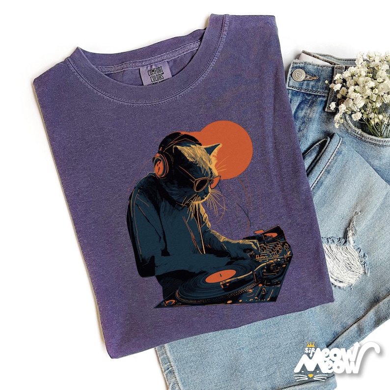 K&ouml;nnte beinhalten: Lila Comfort Colors T-Shirt mit einem DJ-Katzen-Motiv. Die Katze tr&auml;gt Kopfh&ouml;rer und Sonnenbrille und bedient einen Plattenspieler mit einer orangefarbenen Sonne im Hintergrund. Das Shirt wird mit Jeansshorts kombiniert.