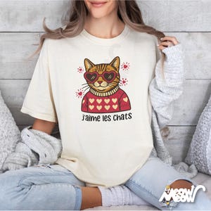 May include: Cream-colored t-shirt featuring a cartoon cat wearing heart-shaped sunglasses and a red sweater with heart patterns. The French text "j'aime les chats" is printed below the cat illustration. The shirt has short sleeves.
