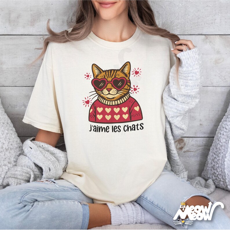 May include: Cream-colored t-shirt featuring a cartoon cat wearing heart-shaped sunglasses and a red sweater with heart patterns. The French text "j'aime les chats" is printed below the cat illustration. The shirt has short sleeves.