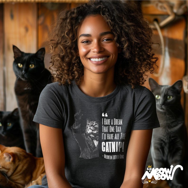 May include: A woman wearing a dark grey t-shirt with a white graphic of a cat holding a paw up in the air. The graphic includes the text "I have a dream that one day I'll have all the catnip! - Meowtin Luther King".