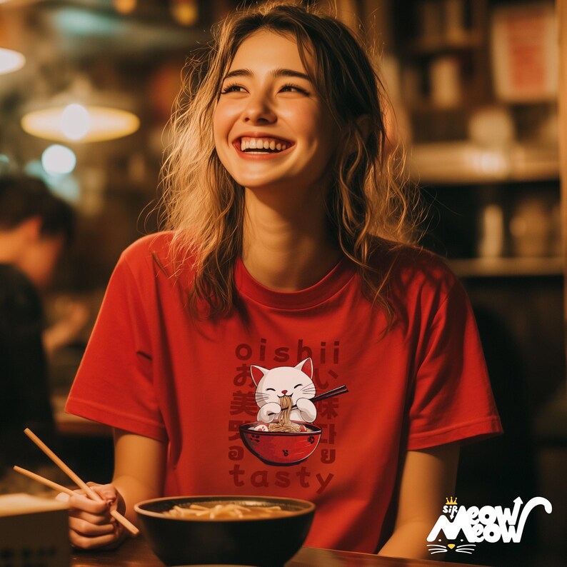 May include: Red t-shirt with a graphic of a white cat eating ramen from a red bowl, with the words "oishii" and "tasty" in Japanese and English. The shirt also has the logo "Meow Meow" in the bottom right corner.