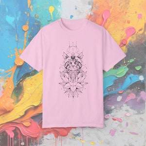 May include: A light pink t-shirt featuring a black graphic of a cat's face above a lotus flower. The cat design has geometric details. The t-shirt is made of a soft material and has short sleeves.