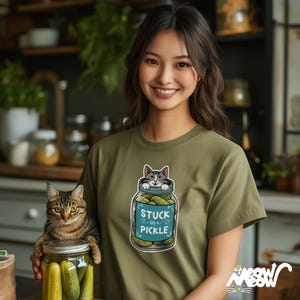 Pu&ograve; includere: Una donna indossa una maglietta verde con una grafica di un gatto bloccato in un barattolo di sottaceti. Il testo sulla grafica dice "Stuck in a Pickle".