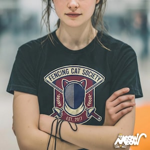 May include: Black t-shirt featuring the text "FENCING CAT SOCIETY" above a fencing mask design, crossed swords, and the text "EST. 2017". The shirt also has a small logo of a cat wearing a crown.