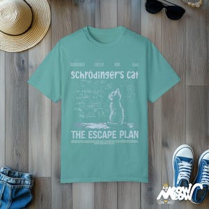 May include: A teal blue t-shirt with a white graphic of a cat looking up at a chalkboard with equations and the text "Schr&ouml;dinger's cat The Escape Plan".