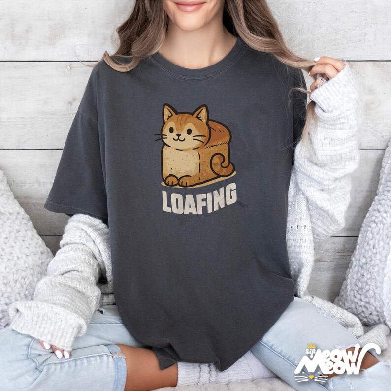 Op de afbeelding: Een donkergrijs T-shirt met een cartoon afbeelding van een kat die in een snee brood ligt. De tekst "LOAFING" staat onder de afbeelding.