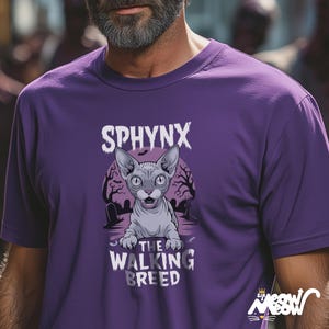 以下が含まれることがあります： 紫色のTシャツ。スフィンクス猫のグラフィックが特徴で、猫の上に「SPHYNX」、下に「THE WALKING BREED」と書かれています。猫は大きな耳を持つグレーで、不気味な墓地のシーンが背景に描かれています。
