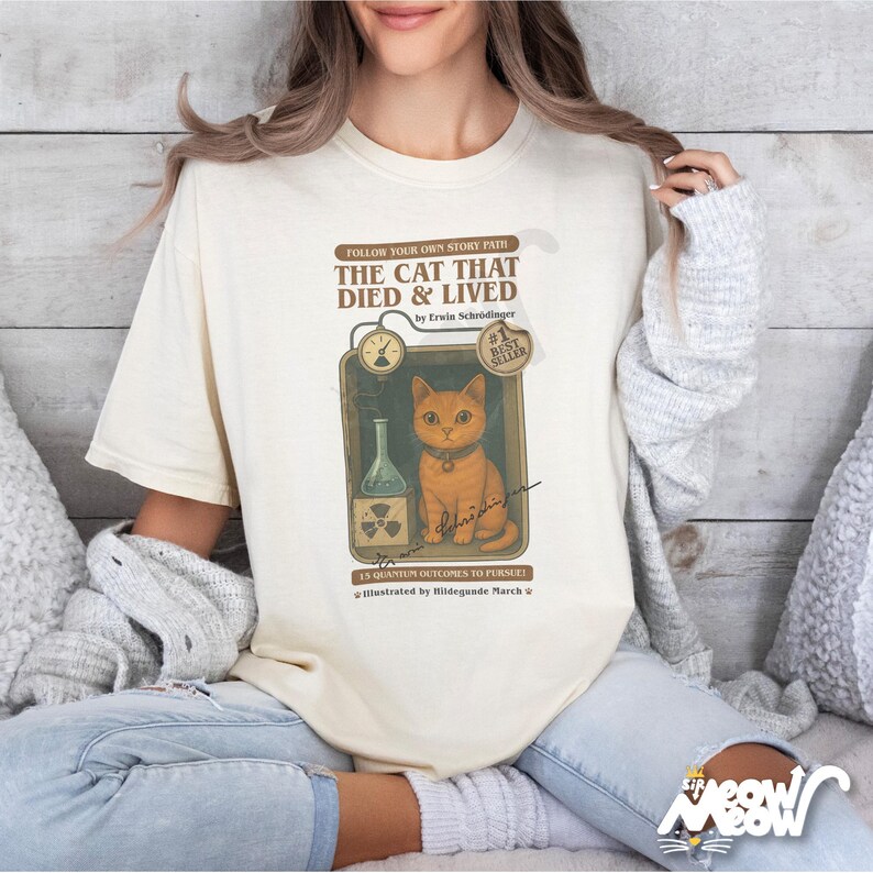 Pu&ograve; includere: Una t-shirt color crema con un design di copertina di libro vintage che presenta un gatto dei cartoni animati in un laboratorio scientifico. Il testo sulla copertina recita "The Cat That Died & Lived" di Erwin Schr&ouml;dinger. Il libro &egrave; etichettato come il #1 Best Seller.