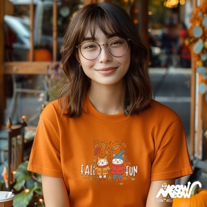 K&ouml;nnte beinhalten: Ein orangefarbenes T-Shirt mit der Aufschrift "FALL FUN" und einer Grafik von zwei Cartoon-Katzen mit M&auml;nteln und H&uuml;ten, umgeben von Herbstbl&auml;ttern. Das T-Shirt wird von einer Person mit braunen Haaren und Brille getragen.