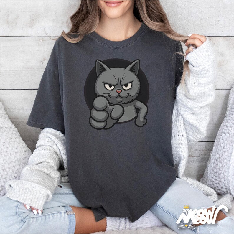 May include: Dark grey t-shirt featuring a cartoon cat with an angry expression, extending a fist. The cat is inside a black circle. The text "Sif Meow" is at the bottom right corner of the shirt.