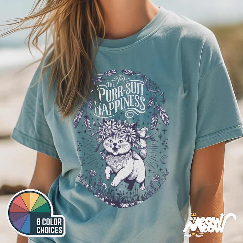 Puede incluir: Una camiseta azul claro con un gr&aacute;fico de un gato con una corona de flores. El texto "The Purr-suit of Happiness" est&aacute; impreso en la camiseta.