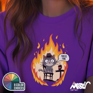 Op de afbeelding: Paars T-shirt met een cartoonkat die een hoed draagt en op een stoel zit, omringd door vlammen. De kat houdt een kopje koffie vast en een spraakballon boven de kat zegt "Dit is prima".