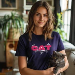 May include: Navy blue t-shirt with the text "CAT mama" in pink and white. The shirt features a cat-themed design. A gray cat is being held in the arms of a person.