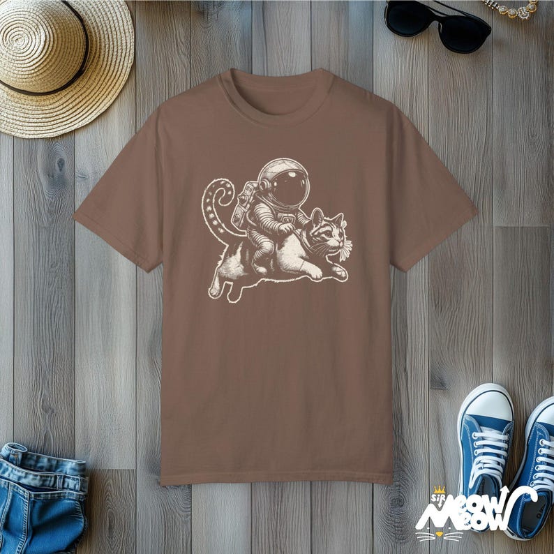 Espresso Comfort Colors 1717 unisex T-shirt with a humorous white graphic of an astronaut riding atop a playful cat, combining outer space and cat lover themes.