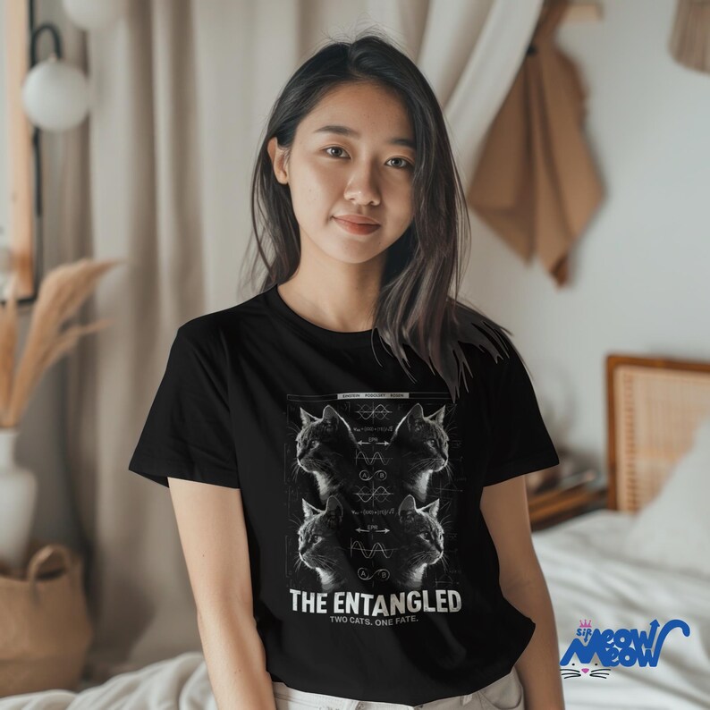 May include: Black t-shirt featuring a graphic of four cat profiles with scientific diagrams and the text "THE ENTANGLED TWO CATS, ONE FATE." The shirt is worn by a person.