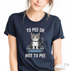 May include: Navy blue t-shirt featuring the text "TO PEE OR NOT TO PEE" with a cartoon cat in a litter box. The cat is grey and white with yellow eyes. The shirt has short sleeves.