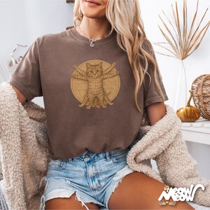 Puede incluir: Una camiseta marr&oacute;n con un gr&aacute;fico de un gato al estilo del Hombre de Vitruvio. El gato es marr&oacute;n y tiene cuatro patas y cuatro brazos extendidos.