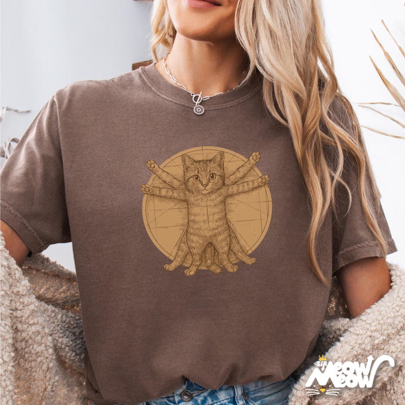 Puede incluir: Una camiseta marr&oacute;n con un gr&aacute;fico de un gato al estilo del Hombre de Vitruvio. El gato tiene cuatro brazos y cuatro patas, y est&aacute; rodeado de un c&iacute;rculo con l&iacute;neas que irradian desde el centro.