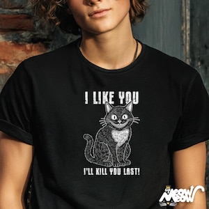 May include: Black t-shirt with a graphic of a smiling black cat with white chest fur. The text "I LIKE YOU" is above the cat, and "I'LL KILL YOU LAST!" is below. The shirt has rolled-up sleeves.