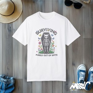 May include: White t-shirt with a black and white illustration of a cat skeleton in a coffin. The text "SURVIVING PURELY OUT OF SPITE" is printed below the image.