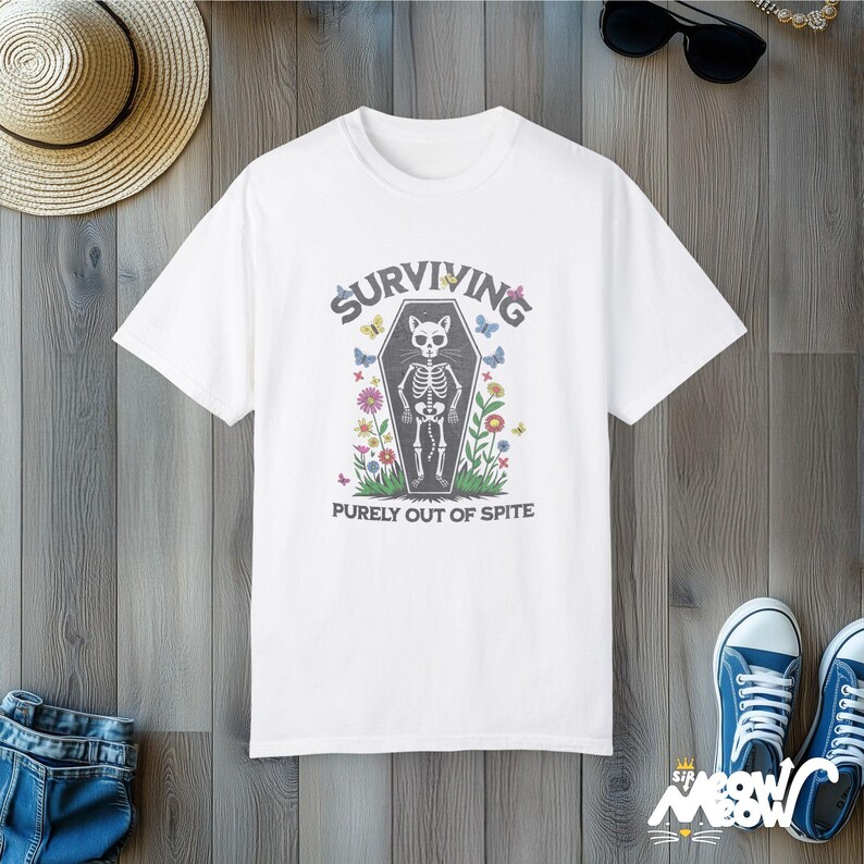 May include: White t-shirt with a black and white illustration of a cat skeleton in a coffin. The text "SURVIVING PURELY OUT OF SPITE" is printed below the image.