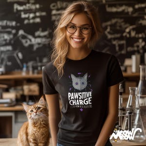 Puede incluir: Una mujer lleva una camiseta negra con un gr&aacute;fico de la cara de un gato y el texto "Pawsitive Charge" y "Electro-Meow-Netic Field of CUDS". Un gato jengibre est&aacute; sentado a su lado.